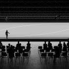 Mockup of an event staging area with an audience, a speaker on stage, and a large screen with minimalistic design. Ideal for graphic designers and event planners.