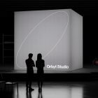 Silhouettes of two people stand in front of a large illuminated display cube with Orbyt Studio logo, suitable for billboard mockups and graphics for designers.
