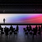 A digital mockup of a presentation event with an audience. It features a wide gradient screen, silhouettes of attendees, ideal for tech or graphics mockups.