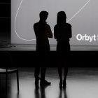 Silhouettes of two people standing in front of a large screen displaying Orbyt Studio. Ideal for print mockups, tech mockups, and graphic design presentations.