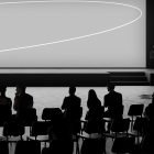 Minimalist auditorium interior scene with silhouettes of people sitting and large screen on stage. Suitable for print mockups, tech mockups, and billboard mockups.