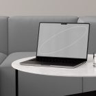 Modern tech mockup showing a laptop on a marble table with a glass of water, situated in a minimalist living space with a grey sofa background.