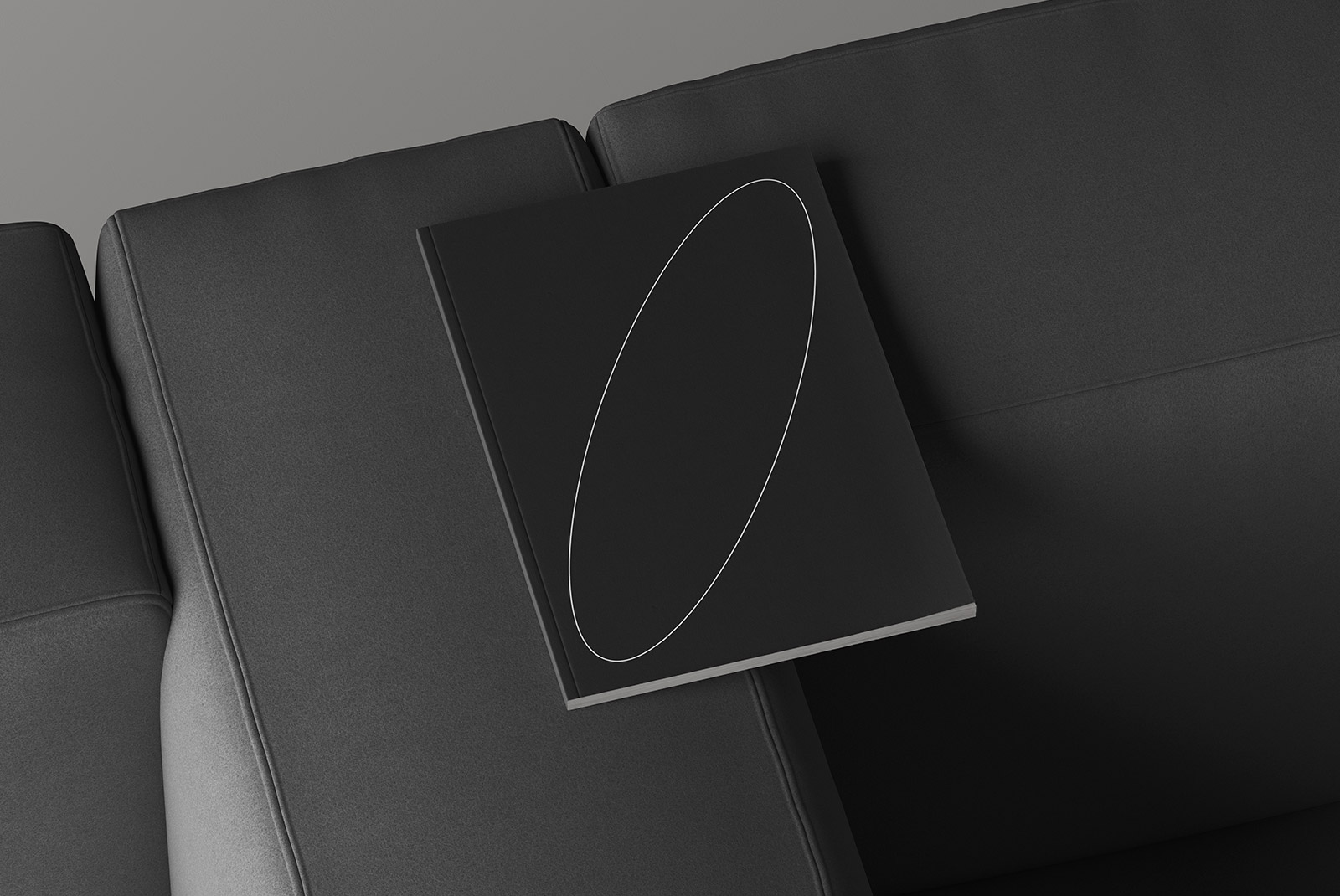 Top view of a minimalistic black hardcover book mockup with a white elliptical design, resting on a gray sofa. Ideal for print and graphic design projects.